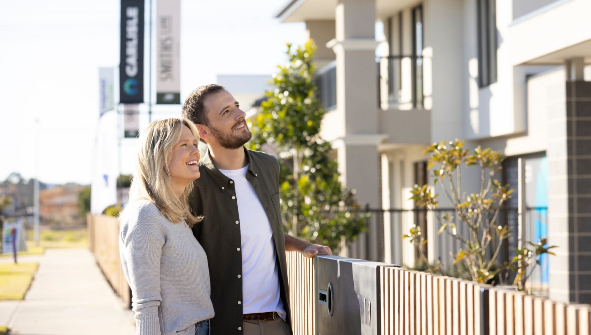Couple smiling by a fence in front of modern apartments.